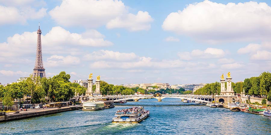 View of the River Seine flowing through Paris, with iconic landmark the Eiffel Tower on the left and historic bridge going across the river. Boats cruise along the water, with blue skies with clouds. 
